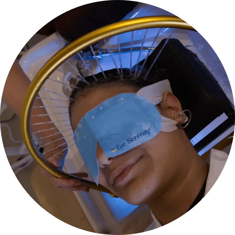 Woman reclined at the shampoo basin wearing a blue eye mask while a gold ring sprays fine streams of water across her scalp